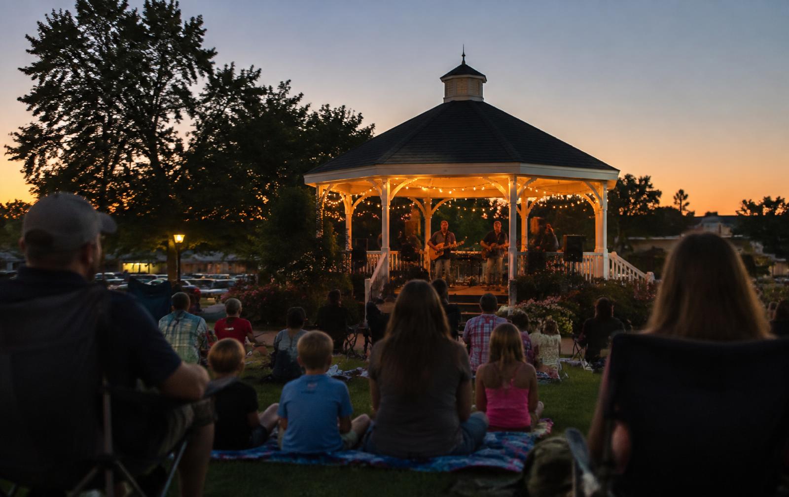 Families on blankets at a Friday evening concert under a string-lit gazebo in a Belmont park